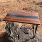Handmade walnut epoxy river accent table with black resin center and black metal hairpin legs, photographed outdoors.